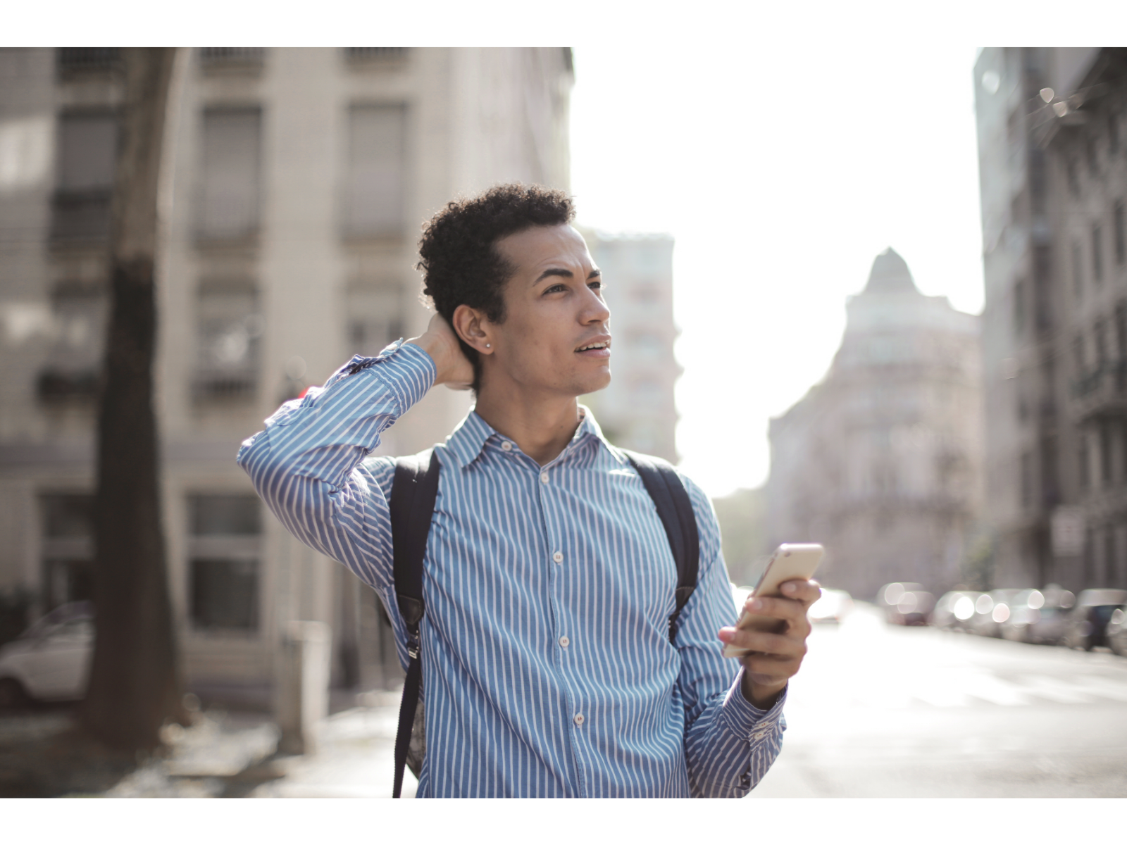 Mildly confused man stands on a city sidewalk and looks off in the distance while holding a smartphone.